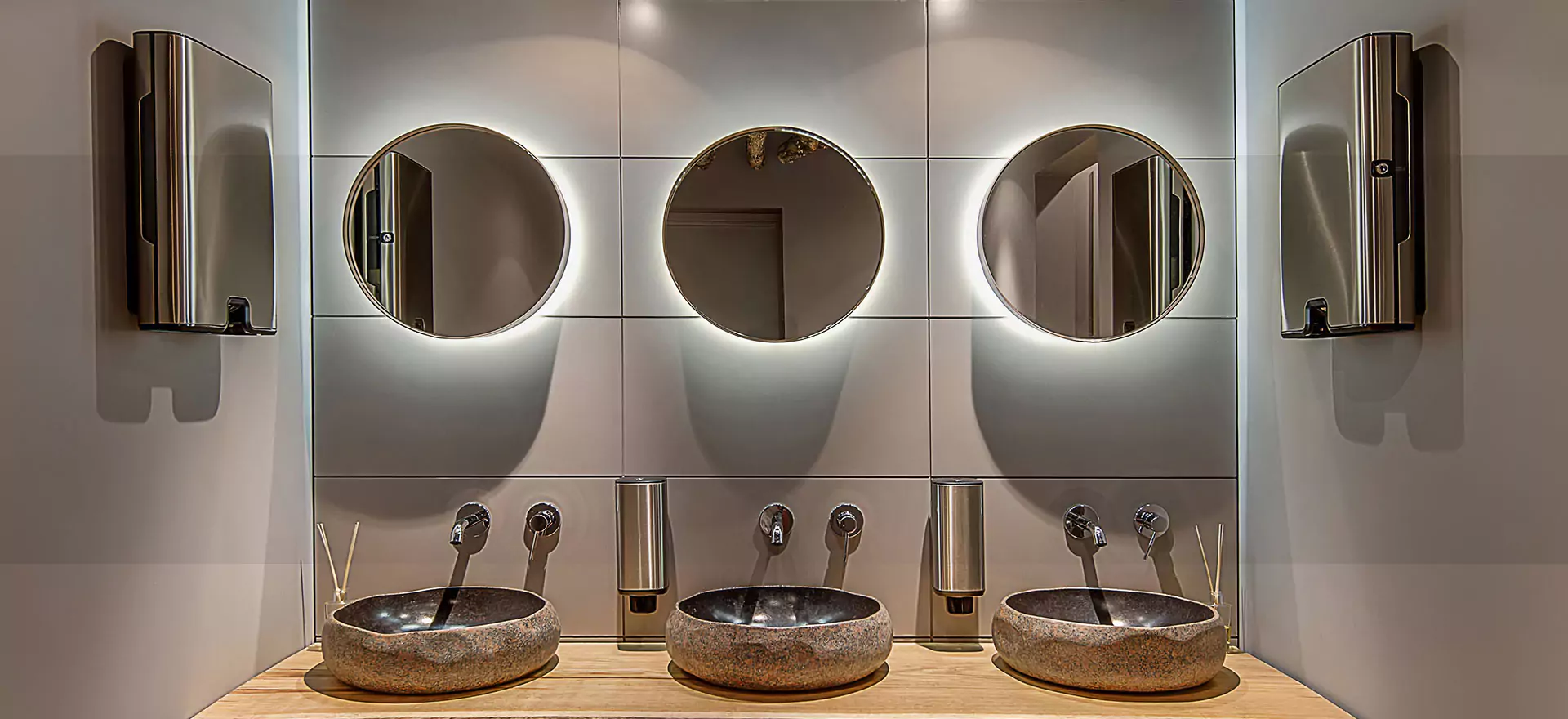 Three circular mirrors hang on a brown wall above the three bathroom sinks.